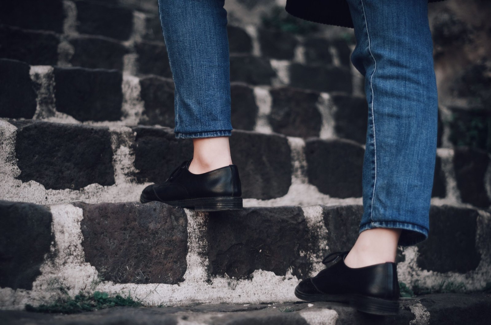 Person in denim jeans climbing stone stairs wearing black leather shoes.